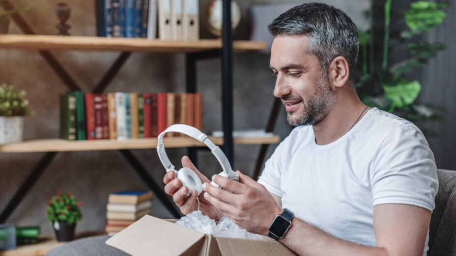 Middle-aged white man sits in front of a bookcase looking at new over-the-headphones as he takes them out of the box.